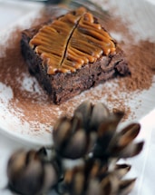 A rich, chocolate brownie topped with a caramel drizzle shaped into a leaf design is centered on a white plate dusted with cocoa powder. In the foreground, there are blurred, artichoke-like decorations.