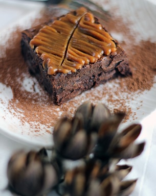 A rich, chocolate brownie topped with a caramel drizzle shaped into a leaf design is centered on a white plate dusted with cocoa powder. In the foreground, there are blurred, artichoke-like decorations.