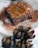 A rich, chocolate brownie topped with a caramel drizzle shaped into a leaf design is centered on a white plate dusted with cocoa powder. In the foreground, there are blurred, artichoke-like decorations.