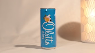 A blue can with Korean text and a cartoon character on it is placed on a light-colored surface. Next to it is a cylindrical object with a textured, honeycomb-like pattern emitting a warm glow.