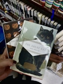 A hand holds two books with decorative covers. One book features a black cat with bright eyes on the cover under the title 'The Great Cat: Poems about Cats', while the other book has a pattern of pumpkin faces. The background consists of shelves filled with books and colorful ribbon bookmarks visible on the right.