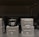 A black scented candle labeled 'Cachemire Bourbon' is placed among several black boxes labeled 'Scented Candle' and 'Oriental Woody.' The boxes and candle are displayed on a dark shelf, with the candle in the center.