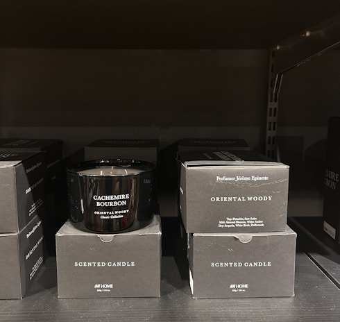 A black scented candle labeled 'Cachemire Bourbon' is placed among several black boxes labeled 'Scented Candle' and 'Oriental Woody.' The boxes and candle are displayed on a dark shelf, with the candle in the center.