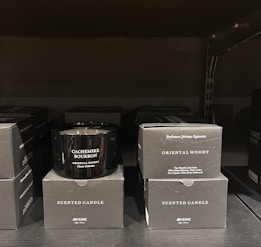 A black scented candle labeled 'Cachemire Bourbon' is placed among several black boxes labeled 'Scented Candle' and 'Oriental Woody.' The boxes and candle are displayed on a dark shelf, with the candle in the center.