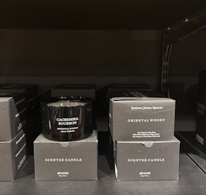 A black scented candle labeled 'Cachemire Bourbon' is placed among several black boxes labeled 'Scented Candle' and 'Oriental Woody.' The boxes and candle are displayed on a dark shelf, with the candle in the center.