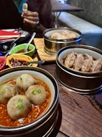A table setting with various dim sum dishes in steamers, including dumplings in a red sauce with green garnish. Accompanying the dishes are colorful bowls containing sauces, and chopsticks are laid out. A person is partially visible in the background holding a small cup.