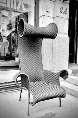 An ornate, high-backed chair with a distinctive woven pattern is positioned on a city sidewalk. Its unique design features rolled ends on both the headrest and armrests, and it is crafted from a dark, sturdy material. The chair stands out against a backdrop of stone building facades and a large window displaying interior plants and decor.