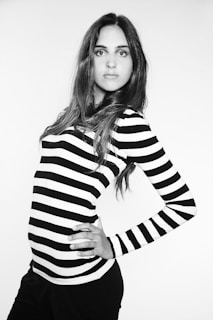 A person with long hair poses confidently in a black and white striped top. The monochrome setting gives a classic and fashionable look. The individual has one hand on their hip, exuding confidence and elegance.