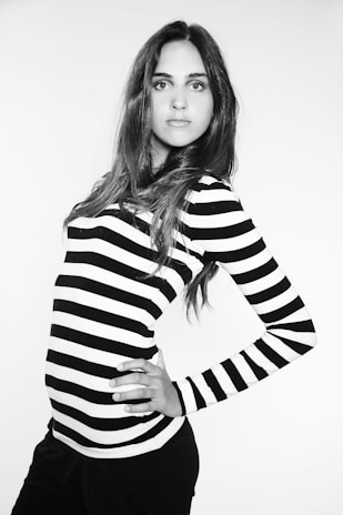 A person with long hair poses confidently in a black and white striped top. The monochrome setting gives a classic and fashionable look. The individual has one hand on their hip, exuding confidence and elegance.