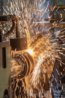A close-up of sparks flying as a metalwork process is taking place. The image captures the vibrant movement of the bright, fiery sparks against a backdrop of machinery parts. There is a sense of intense energy and industrial activity.