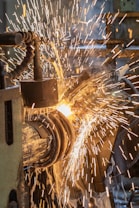 A close-up of sparks flying as a metalwork process is taking place. The image captures the vibrant movement of the bright, fiery sparks against a backdrop of machinery parts. There is a sense of intense energy and industrial activity.