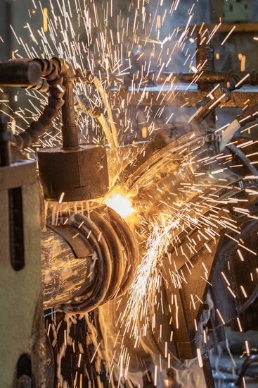 A close-up of sparks flying as a metalwork process is taking place. The image captures the vibrant movement of the bright, fiery sparks against a backdrop of machinery parts. There is a sense of intense energy and industrial activity.