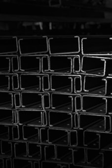 A black and white photo of stacked metal beams with a distinct 'I' shape, showcasing the industrial aesthetic and geometry.