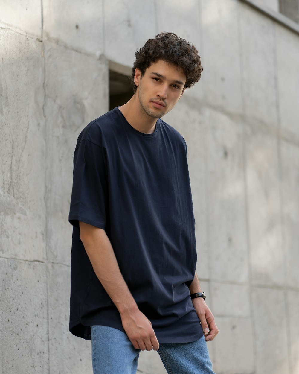 a man in a black t - shirt and blue jeans is leaning against a wall