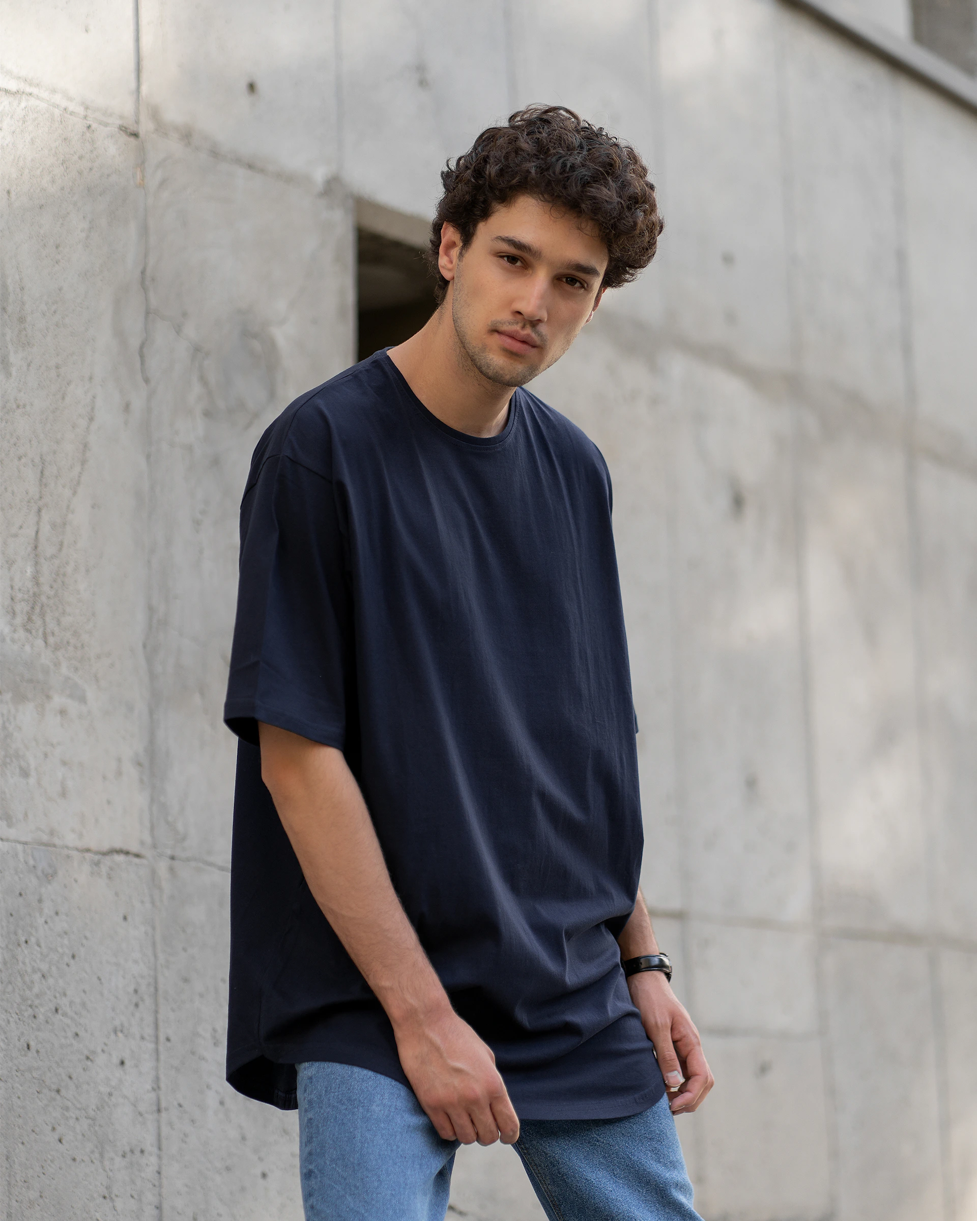 a man in a black t - shirt and blue jeans is leaning against a wall