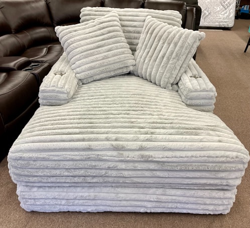 A large, plush, cushioned piece of furniture with a soft, quilted texture, featuring horizontal stripes. It includes two matching throw pillows placed on top, positioned in a well-lit room with a brown leather recliner and other furniture visible in the background.