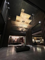 A modern interior space with an artistic ceiling structure made of layered, golden panels. The walls feature vertical stripes and there is a large, square coffee table centered in the open area. Dim lighting and elegant furniture create a luxurious atmosphere.