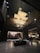 A modern interior space with an artistic ceiling structure made of layered, golden panels. The walls feature vertical stripes and there is a large, square coffee table centered in the open area. Dim lighting and elegant furniture create a luxurious atmosphere.