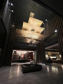 A modern interior space with an artistic ceiling structure made of layered, golden panels. The walls feature vertical stripes and there is a large, square coffee table centered in the open area. Dim lighting and elegant furniture create a luxurious atmosphere.