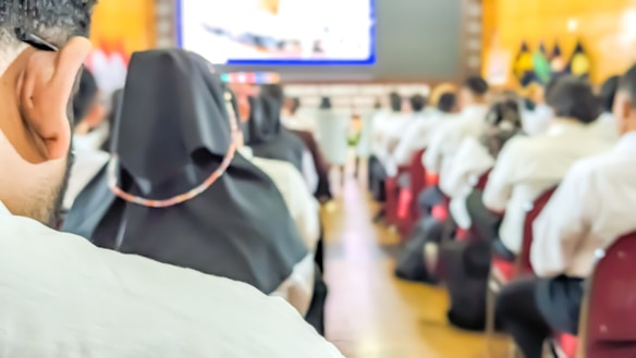 A conference or seminar setting with people seated in rows, facing a large screen displaying a presentation. Many attendees are in formal attire with some wearing head coverings.