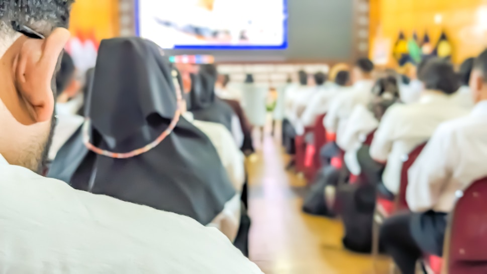 A conference or seminar setting with people seated in rows, facing a large screen displaying a presentation. Many attendees are in formal attire with some wearing head coverings.