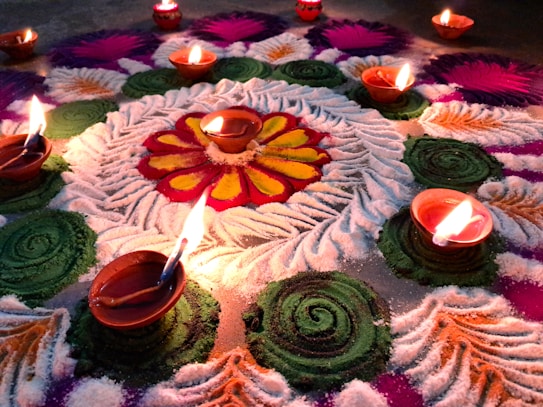 An elaborate and colorful rangoli design created with vibrant powders is accented by small clay oil lamps with flickering flames. The rangoli features intricate swirls and floral patterns in shades of green, pink, purple, white, and red. The central motif resembles a bright flower surrounded by leaf-like patterns.