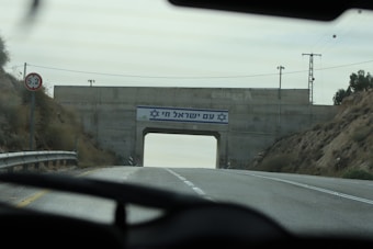 A road passes under a concrete bridge with a sign featuring Hebrew text and a blue Star of David. The surrounding area includes hilly terrain with sparse vegetation. A road sign indicates a height clearance of 5.2 meters, and there are road markings and guardrails visible.