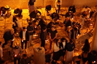 People are gathered, many kneeling on stone pavement while holding candles. The warm glow of the candles creates a serene and solemn atmosphere, with the light casting soft shadows on the ground. Some individuals are conversing or praying.