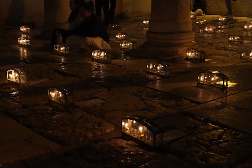 Lanterns with candles softly illuminate a textured cobblestone pavement. Several people are seated around these lanterns in a dimly lit, possibly outdoor space. Stone columns are visible in the background.
