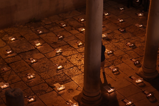 A shadowed temple courtyard at dusk, with a lone girl standing watchful among ancient stone lanterns.