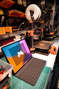 A workspace featuring a 3D printer and a tablet with a keyboard. The 3D printer, equipped with a spool of filament, is actively printing an object with orange components. Nearby, a tablet with a colorful screen is positioned on a cutting mat. The background includes shelves with various tools and supplies, indicating an organized and tech-oriented setting.