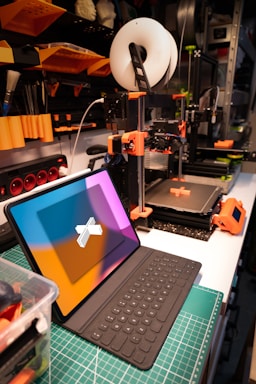 A workspace featuring a 3D printer and a tablet with a keyboard. The 3D printer, equipped with a spool of filament, is actively printing an object with orange components. Nearby, a tablet with a colorful screen is positioned on a cutting mat. The background includes shelves with various tools and supplies, indicating an organized and tech-oriented setting.