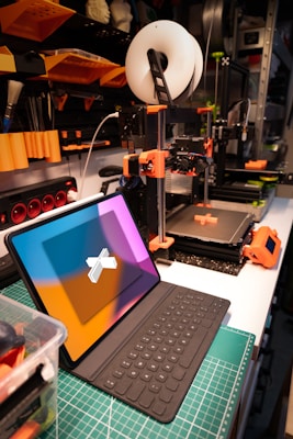 A workspace featuring a 3D printer and a tablet with a keyboard. The 3D printer, equipped with a spool of filament, is actively printing an object with orange components. Nearby, a tablet with a colorful screen is positioned on a cutting mat. The background includes shelves with various tools and supplies, indicating an organized and tech-oriented setting.
