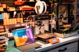 A well-organized workshop with 3D printers, tools, and a tablet. The workspace includes two 3D printers on a workbench, surrounded by tools like paintbrushes and pencils neatly arranged on a pegboard. Bright orange and black are predominant, with a laptop displaying a colorful screen in the foreground.