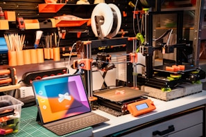 A well-organized workshop with 3D printers, tools, and a tablet. The workspace includes two 3D printers on a workbench, surrounded by tools like paintbrushes and pencils neatly arranged on a pegboard. Bright orange and black are predominant, with a laptop displaying a colorful screen in the foreground.