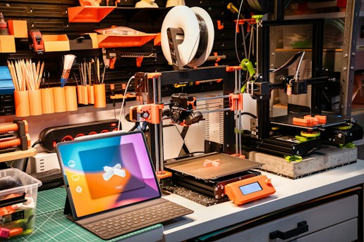 A well-organized workshop with 3D printers, tools, and a tablet. The workspace includes two 3D printers on a workbench, surrounded by tools like paintbrushes and pencils neatly arranged on a pegboard. Bright orange and black are predominant, with a laptop displaying a colorful screen in the foreground.