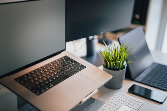 A workspace setup featuring a laptop with its screen slightly open, a tablet, a smartphone, and a wireless keyboard on a desk. A potted green plant adds a touch of nature to the sleek and modern tech environment. Multi-screen setup suggests a focus on productivity.