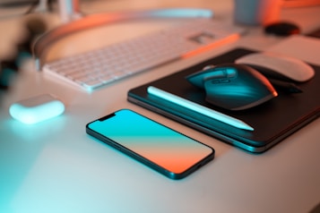 A modern desk setup featuring a smartphone with a blue and orange gradient screen, a sleek wireless mouse, a wireless keyboard, and a stylus on top of a tablet. An open case for wireless earbuds is also visible, all arranged on a clean white surface with ambient lighting.