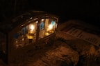 Two lit candles are enclosed in a small glass and metal case with blue decorations including heart shapes and stars. The case is situated on a stone surface with a pamphlet or flyer partially visible in the background.