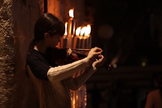 A person is playing a violin against a dimly-lit background with several lit candles nearby. The warm glow from the candles casts a soft light on the musician, creating a serene and intimate atmosphere.