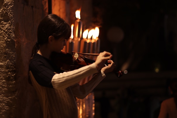 A person is playing a violin against a dimly-lit background with several lit candles nearby. The warm glow from the candles casts a soft light on the musician, creating a serene and intimate atmosphere.