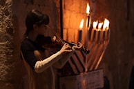 A person plays the violin next to a lit menorah with seven visible burning candles, set against a textured stone wall. The warm glow from the candles illuminates the scene, casting a soft light on the musician.