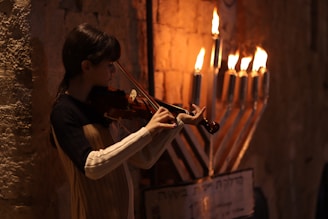 A person plays the violin next to a lit menorah with seven visible burning candles, set against a textured stone wall. The warm glow from the candles illuminates the scene, casting a soft light on the musician.