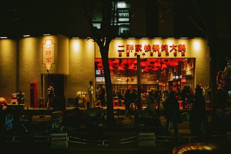 A bustling street scene outside a brightly lit restaurant with red signage and interior. People are gathered inside, likely enjoying a meal, while others stand and move about outside. Red chairs and lanterns contribute to a vibrant atmosphere.