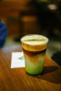 A glass of iced matcha latte with a layered appearance, featuring green matcha at the bottom, white milk in the middle, and a layer of brown coffee on top. The drink is placed on a wooden table.