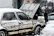 A smiling roadside assistance technician helping a customer beside a car with the hood open on a snowy street.
