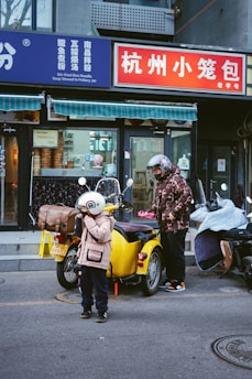 A child and an adult stand next to a yellow motorbike with a sidecar in front of a shop. The shop has signs with both Chinese and English text. The child wears a white helmet and pink coat, while the adult has a floral-patterned coat and helmet. Other scooters are parked nearby.