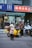 A child and an adult stand next to a yellow motorbike with a sidecar in front of a shop. The shop has signs with both Chinese and English text. The child wears a white helmet and pink coat, while the adult has a floral-patterned coat and helmet. Other scooters are parked nearby.
