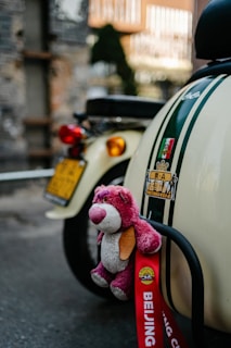 A stuffed pink bear with a beige belly and snout is attached to the back of a cream-colored scooter. The scooter has green racing stripes and various stickers, including one with Asian characters. A red ribbon with the text 'BEIJING' is hanging, indicating a possible location or event. The background is an urban setting with a blurred mix of textures and colors.