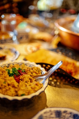 A dish of food is displayed in the foreground on a table. It appears to be garnished with green herbs and red chili peppers. A spoon is placed inside the dish. The background is blurred and includes various other items and kitchen utensils.